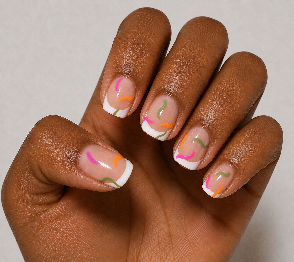 A close-up of a hand with manicured nails featuring a white French tip design and delicate abstract curved lines in pink, orange, and olive green.