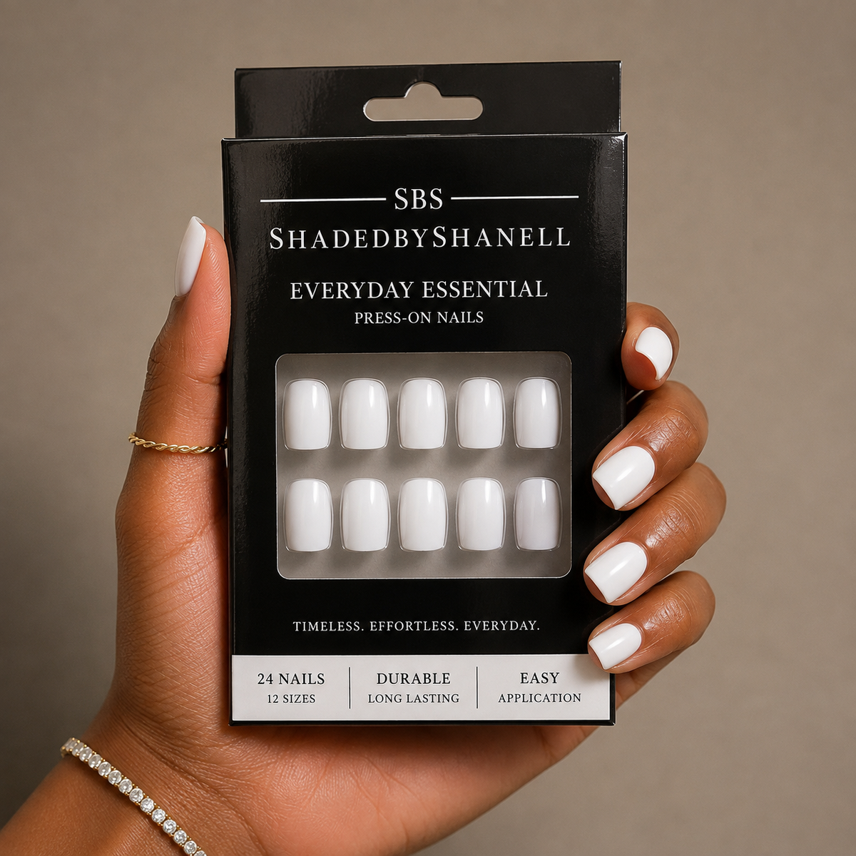 A hand holding a black box of SBS ShadedbyShanell Everyday Essential Press-On Nails. The nails visible through the clear window are white, and the hand has white manicured nails.