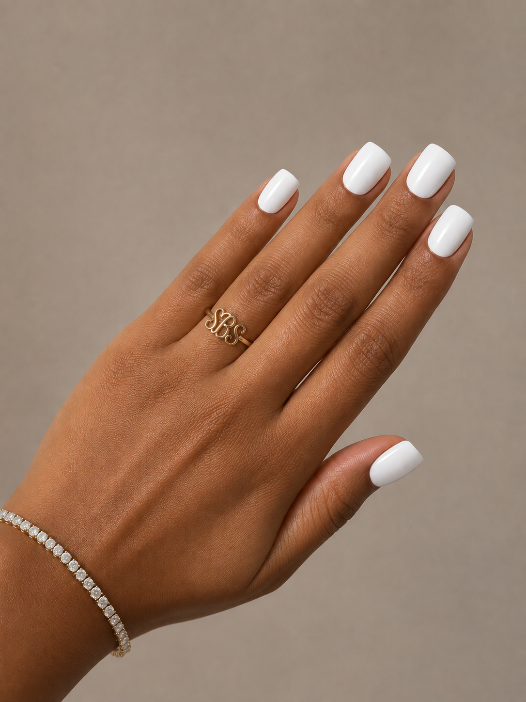 A close-up of a hand with white manicured nails, wearing a delicate gold ring with an intertwined monogram design and a thin diamond-embellished bracelet, set against a solid taupe background.