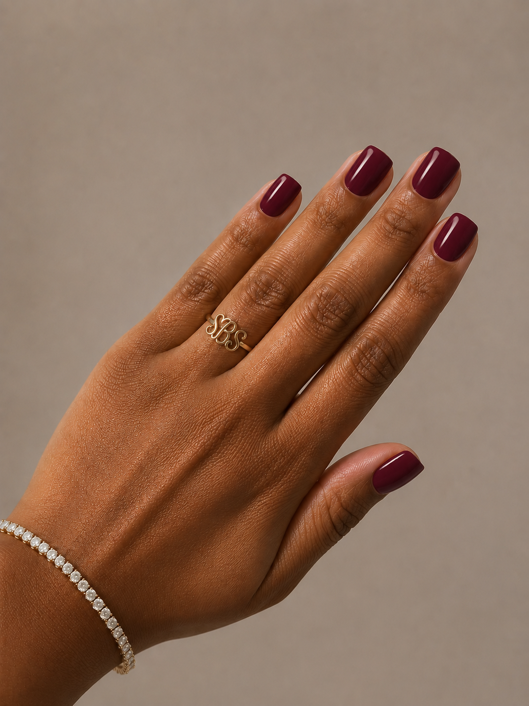 A close-up of a hand with dark burgundy manicured nails, wearing a gold ring with a stylized 'SBS' design and a delicate diamond bracelet, set against a solid taupe background.