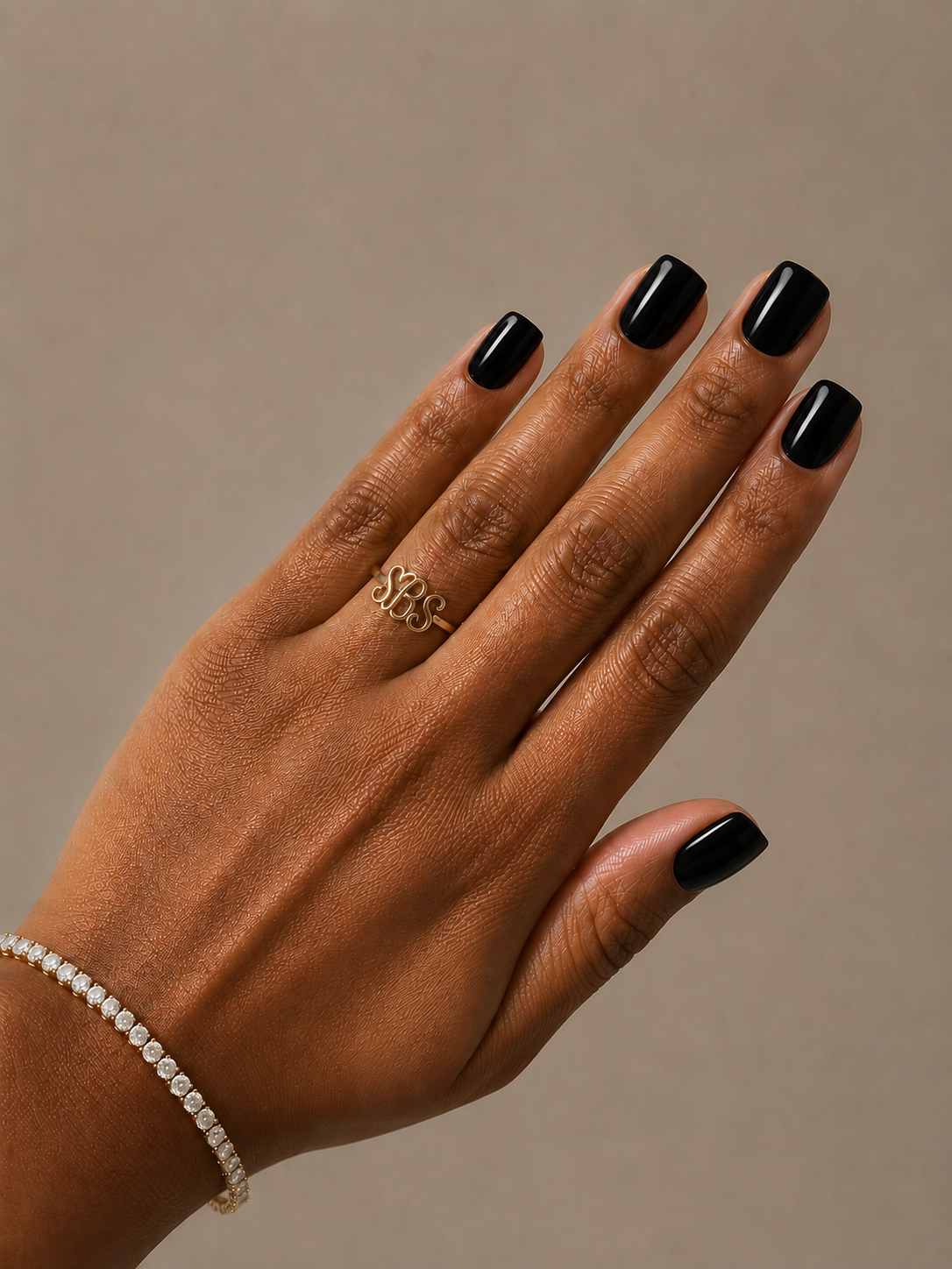 A close-up of a hand with black manicured nails wearing a delicate gold ring with an interlocking monogram design, set against a solid warm taupe background.