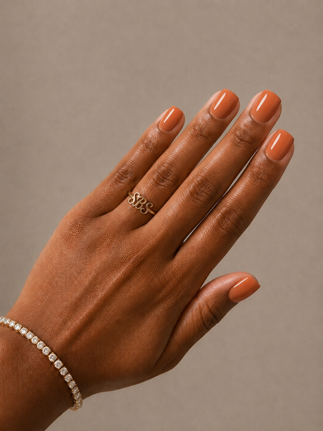 A close-up of a hand with orange manicured nails, wearing a gold ring with the word 'SBS' in cursive and a delicate gold bracelet with clear stones, set against a solid taupe background.
