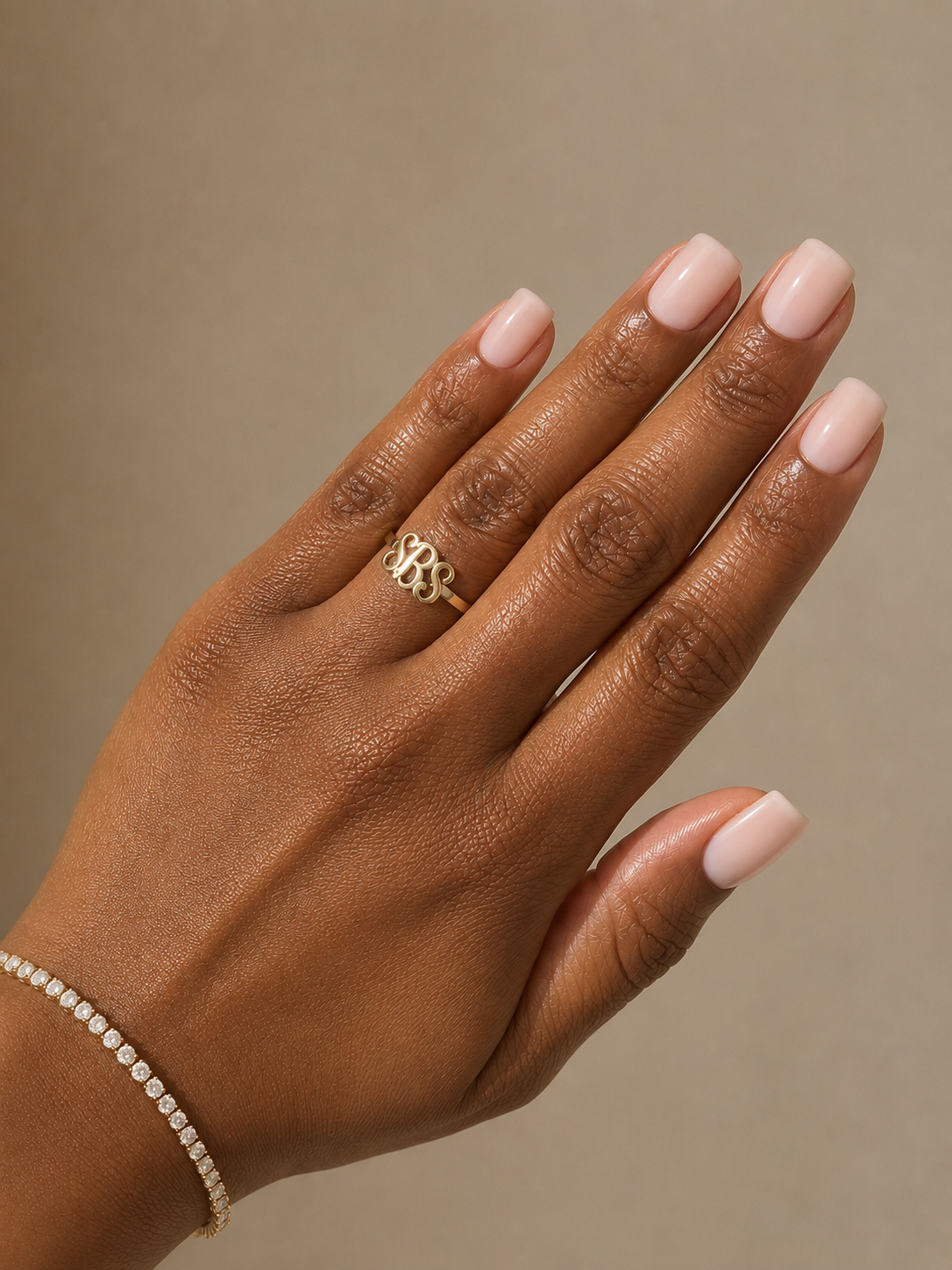 A close-up of a hand with pale pink manicured nails wearing a gold ring with interlocking letters and a delicate gold bracelet against a solid taupe background.