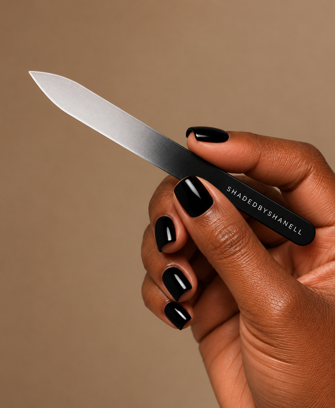A hand with black manicured nails holds a slim black and silver nail file against a solid warm beige background. The handle is printed with the brand name 'SHADEDBYSHANELL'.