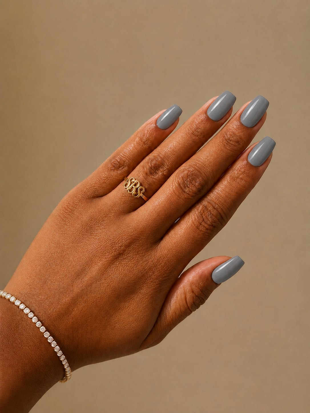 A close-up of a hand with gray-blue manicured nails and a gold ring, shown against a warm beige background. A delicate pearl bracelet is also visible on the wrist.