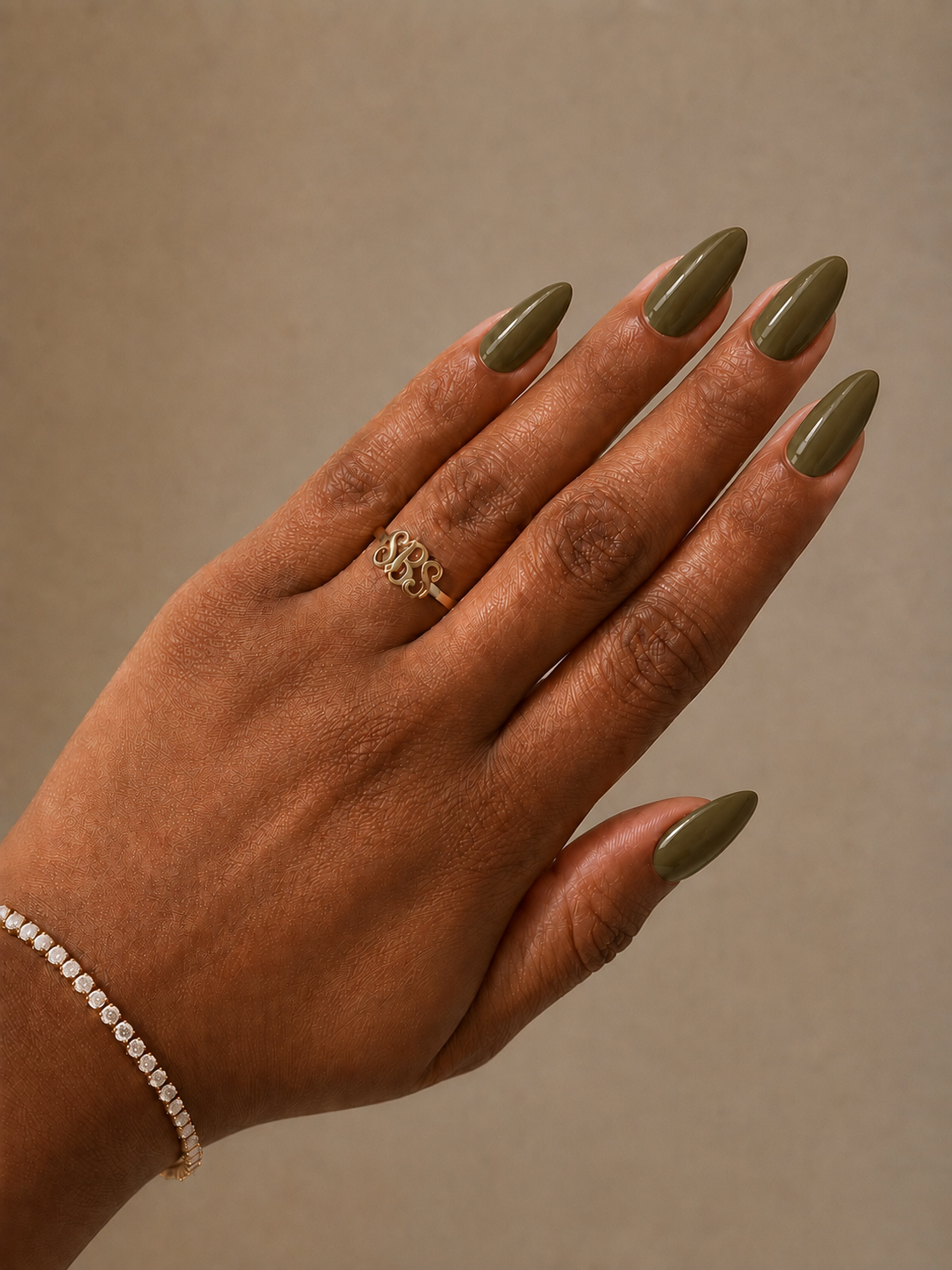 A close-up of a hand with olive green manicured nails wearing a gold ring with an interlocking monogram design, set against a solid beige background.